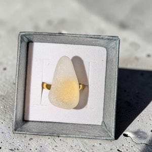 Könnte beinhalten: Ein goldfarbener Ring mit einem glatten, mattierten, tropfenförmigen Stück Meerglas, das ein durchscheinendes weißes Äußeres und ein zartgelbes Zentrum aufweist, präsentiert in einer quadratischen, grauen Schmuckschachtel aus Karton mit weißem Inlay.
