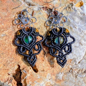 May include: A pair of handmade earrings featuring intricate black macrame designs. Each earring has a central, iridescent green bead and is accented with gold thread. The earrings are suspended from silver-toned wirework and are displayed on a textured, light brown surface.