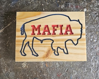 May include: Wooden sign featuring a black outline of a bison with the word "MAFIA" in red letters. The sign is made of light-colored wood and has a rustic appearance.