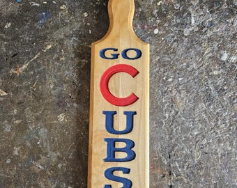 May include: A wooden paddle with the words "GO CUBS GO!" carved and painted in blue and red. The paddle has a handle and a hole for hanging. The background is a mottled, multi-colored surface.