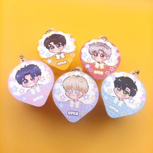 May include: Five heart-shaped acrylic charms featuring anime-style characters with large eyes and bows. Each charm has a metal ball chain, a grid pattern background with stars, and the word OPEN printed in white text on the lower section.