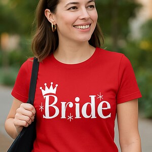 May include: A red short-sleeved t-shirt featuring the white text "Bride" printed on the front. The design includes a white crown graphic above the word and small white starburst accents surrounding the lettering, creating a clear graphic print.