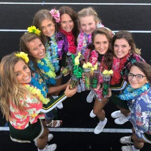 May include: A group of eight young women wearing green and white cheerleading uniforms and colorful leis, holding clear plastic cups with green and yellow contents. They are smiling and looking at the camera.