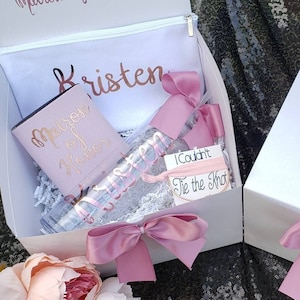 May include: Two white gift boxes with pink satin bows. The box on the left has a white robe, a tumbler with the text "Matron of Honor", a water bottle with the text "Kristen", and a small card with the text "I Couldn't Tie the Knot". The box on the right has the name "Rachel" written on the top.
