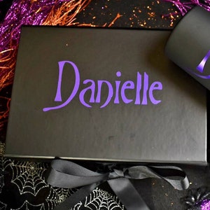 May include: A black gift box and a black tumbler, both with the name "Danielle" in purple script. The tumbler is cylindrical. The background includes black fabric, glitter, and decorative elements in purple and orange.