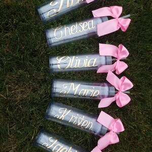 May include: Six clear plastic tumblers with pink bows and straws. Each tumbler has a gold name on it: Jill, Chelsea, Olivia, Marie, Kristen, and Jackie.
