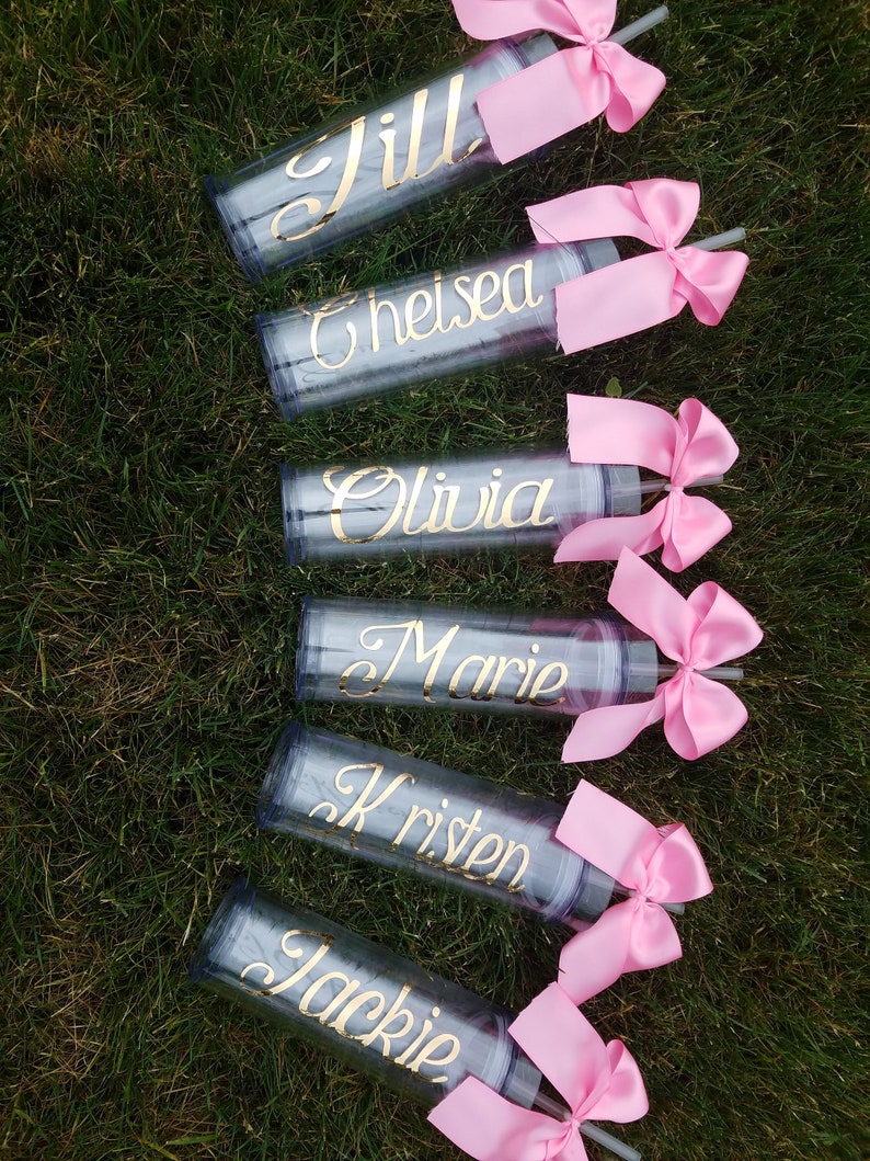 May include: Six clear plastic tumblers with pink bows and straws. Each tumbler has a gold name on it: Jill, Chelsea, Olivia, Marie, Kristen, and Jackie.