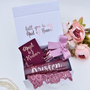 May include: A white gift box with a pink ribbon and a card that says "Will you be my Maid of Honor?" The box contains a burgundy can cooler with gold lettering that says "Maid of Honor", a burgundy tumbler with gold lettering that says "Kristen", a burgundy and pink patterned scarf, and a pink lace doily.