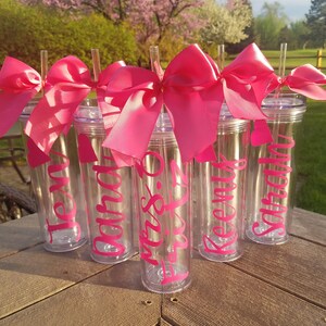 May include: Six clear plastic tumblers with pink bows and straws. Each tumbler has a name written on it in pink lettering: Jen, Sarah,  Miss.  to  be,  Keenan,  and  Sophia.