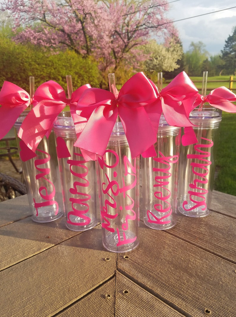 May include: Six clear plastic tumblers with pink bows and straws. Each tumbler has a name written on it in pink lettering: Jen, Sarah,  Miss.  to  be,  Keenan,  and  Sophia.
