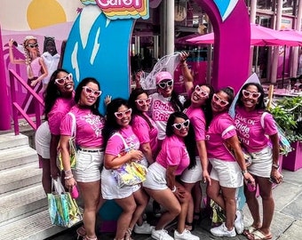 May include: A group of people wearing pink t-shirts, white shorts, and heart-shaped sunglasses pose in front of a pink archway with the text "Malibu Barbie Cafe". The cafe has a pink and blue color scheme.