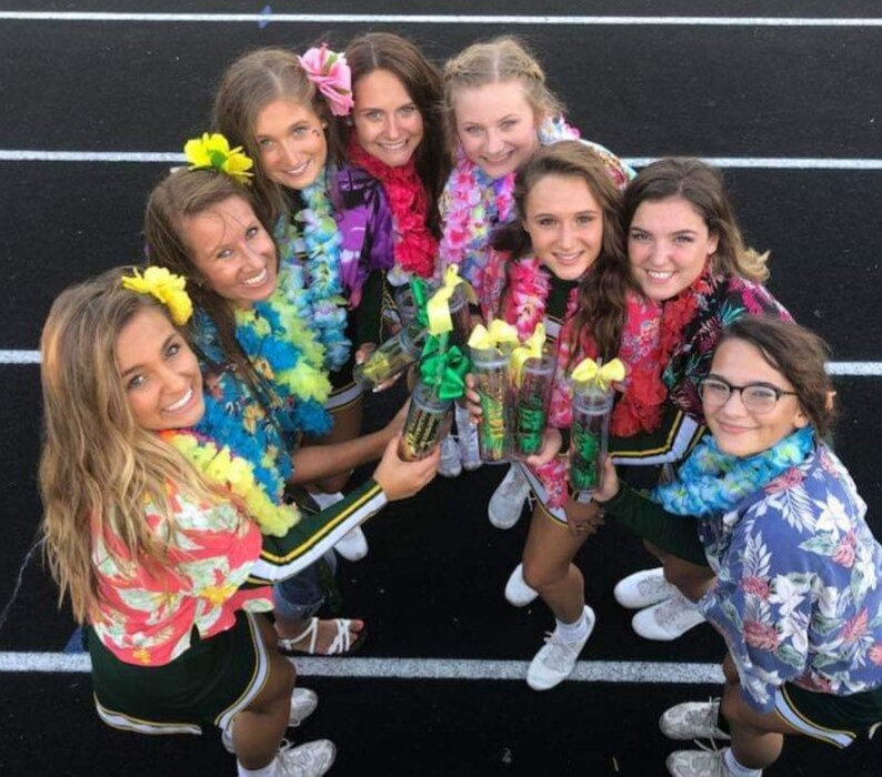 May include: A group of eight young women wearing green and white cheerleading uniforms and colorful leis, holding clear plastic cups with green and yellow contents. They are smiling and looking at the camera.