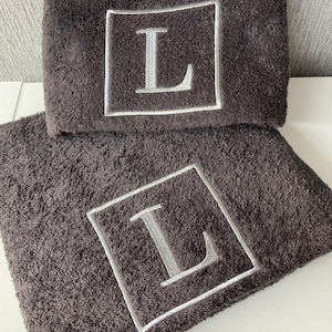 May include: Two dark gray bath towels with a plush texture. Each towel features a large, embroidered letter "L" in silver within a white square frame. The towels are neatly folded and stacked, showcasing the personalized design.