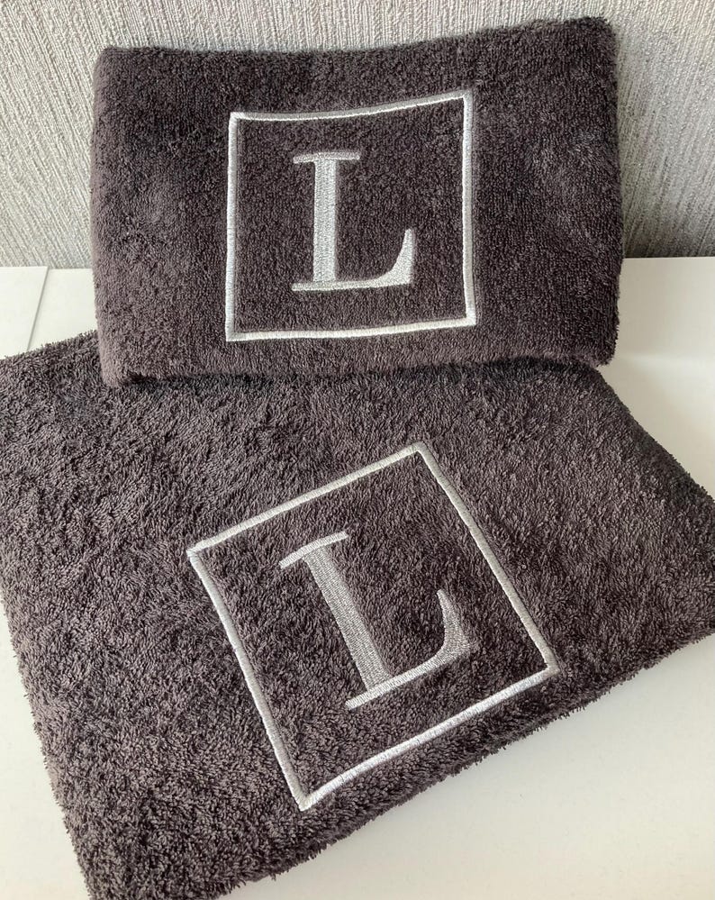 May include: Two dark gray bath towels with a plush texture. Each towel features a large, embroidered letter "L" in silver within a white square frame. The towels are neatly folded and stacked, showcasing the personalized design.