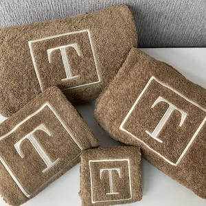 May include: A set of five brown bath towels with the letter "T" embroidered in white. Each towel has a white border around the embroidered letter. The towels are arranged in a neat, organized display.