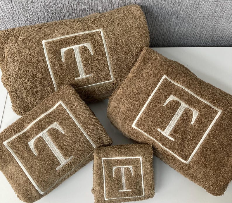 May include: A set of five brown bath towels with the letter "T" embroidered in white. Each towel has a white border around the embroidered letter. The towels are arranged in a neat, organized display.
