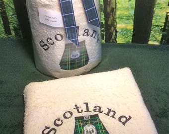 May include: A white bath towel with the word "Scotland" embroidered in black, along with a green and yellow tartan kilt design. A matching towel set is wrapped in clear cellophane and tied with a plaid ribbon.