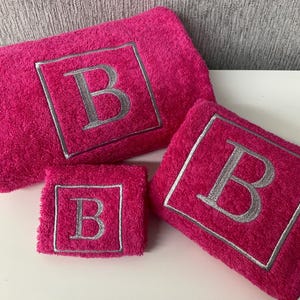 May include: A set of three hot pink terry cloth towels. Each towel features a silver embroidered letter "B" within a square frame. The set includes a large bath towel, a hand towel, and a washcloth.