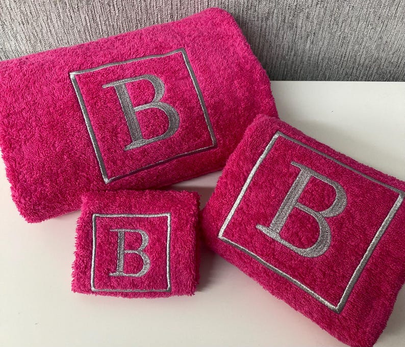 May include: A set of three hot pink terry cloth towels. Each towel features a silver embroidered letter "B" within a square frame. The set includes a large bath towel, a hand towel, and a washcloth.