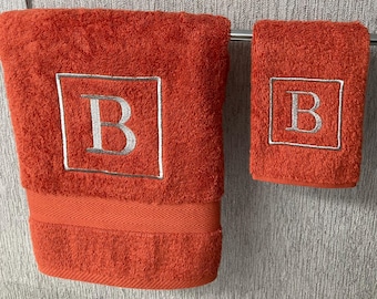 Könnte beinhalten: Zwei orangefarbene Badetücher, eines groß und eines klein, hängen an einer Metallstange. Jedes Handtuch hat den silbernen Buchstaben "B" in einem quadratischen Rahmen. Die Handtücher sind aus einem weichen Material.