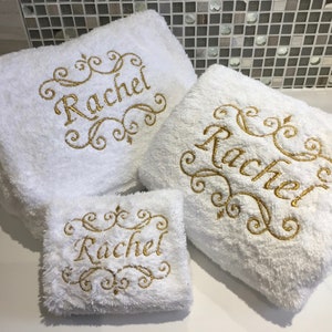 May include: Three white, fluffy towels with gold embroidered text that reads "Rachel". The towels are in different sizes, with decorative scrollwork around the name. The towels are stacked on top of each other.