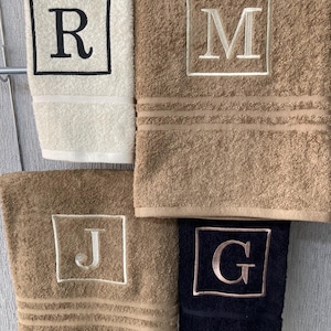 May include: Four plush bath towels in cream, tan, and navy blue, each embroidered with a single initial inside a square frame. The letters are R, M, J, and G. The towels have a textured appearance.