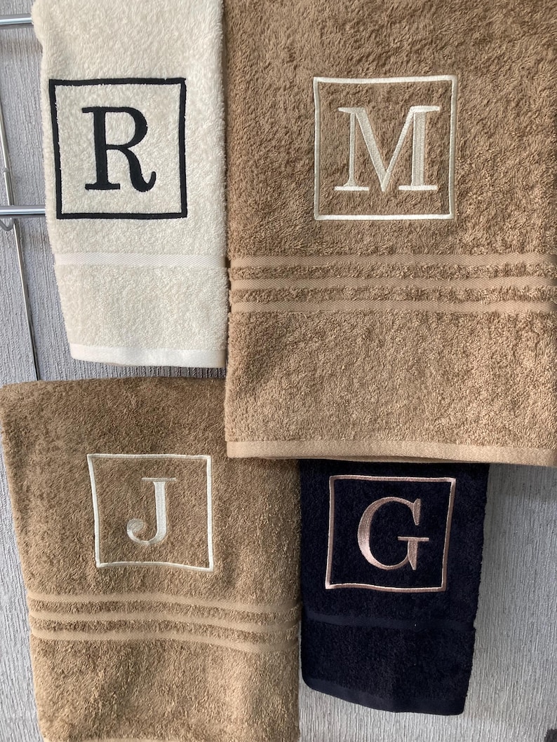 May include: Four plush bath towels in cream, tan, and navy blue, each embroidered with a single initial inside a square frame. The letters are R, M, J, and G. The towels have a textured appearance.