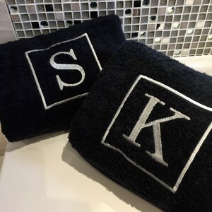 May include: Two black bath towels with embroidered silver letters, "S" and "K", inside white square frames. The towels are plush and folded, suggesting a bathroom or spa setting. The background features a mosaic tile wall.
