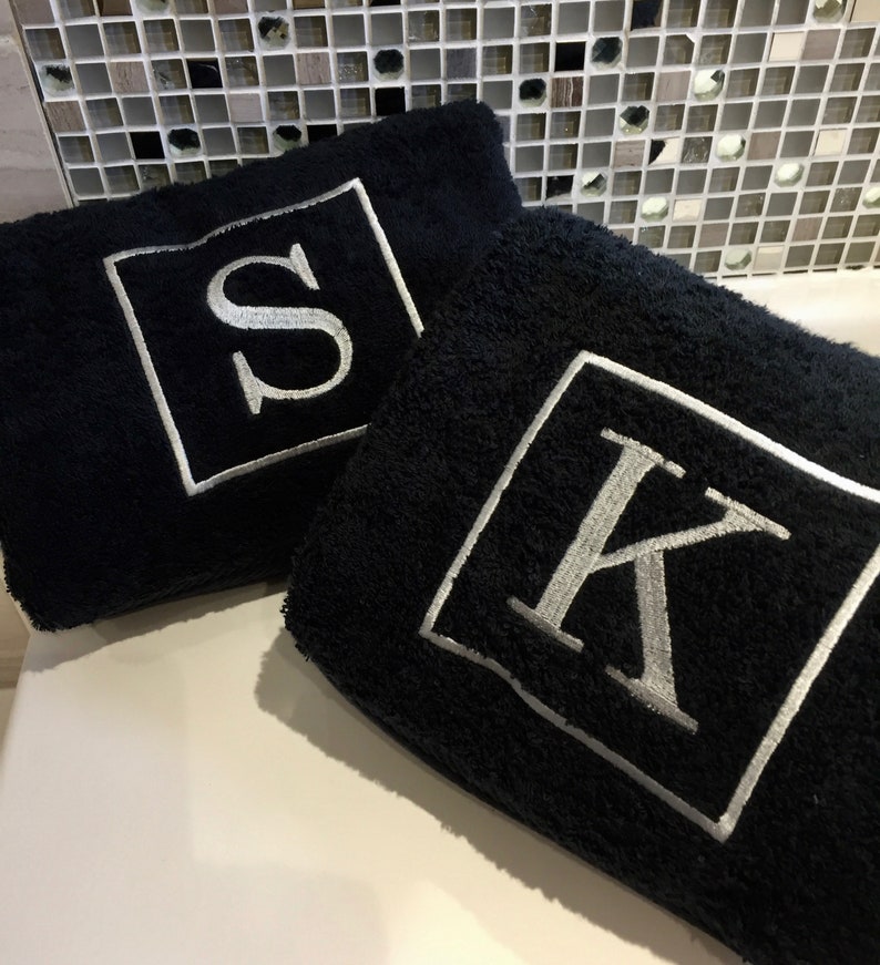 May include: Two black bath towels with embroidered silver letters, "S" and "K", inside white square frames. The towels are plush and folded, suggesting a bathroom or spa setting. The background features a mosaic tile wall.
