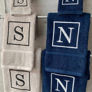 May include: A set of beige and navy blue bath towels, each embroidered with a single initial within a square frame. The towels are hanging on a metal rack, showcasing the contrasting colors and personalized designs.