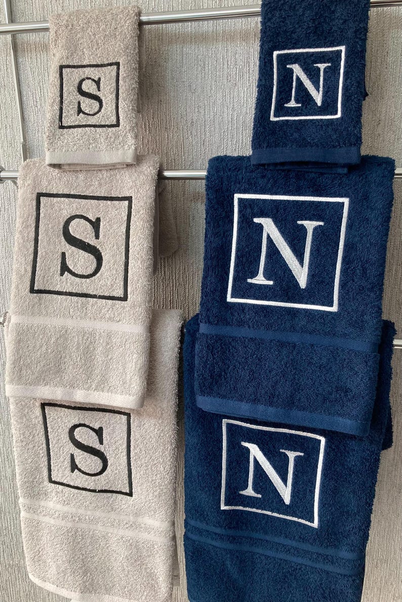 May include: A set of beige and navy blue bath towels, each embroidered with a single initial within a square frame. The towels are hanging on a metal rack, showcasing the contrasting colors and personalized designs.