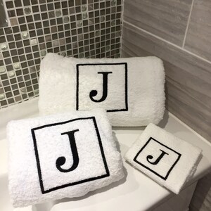May include: Three white terry cloth towels of varying sizes, each embroidered with a black square containing the letter "J". The towels are arranged on a white surface, with a tiled wall in the background.