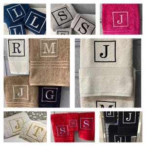 May include: Collection of monogrammed bath towels in various colors, including navy, gray, pink, beige, and red. Each towel features a single initial embroidered in a square frame. The letters include L, S, J, R, M, G, and T. The towels are displayed folded and stacked.