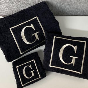 May include: A set of three black terry cloth towels. Each towel features a white embroidered square with the letter "G" in the center. The towels are in varying sizes, suitable for a bathroom or spa.
