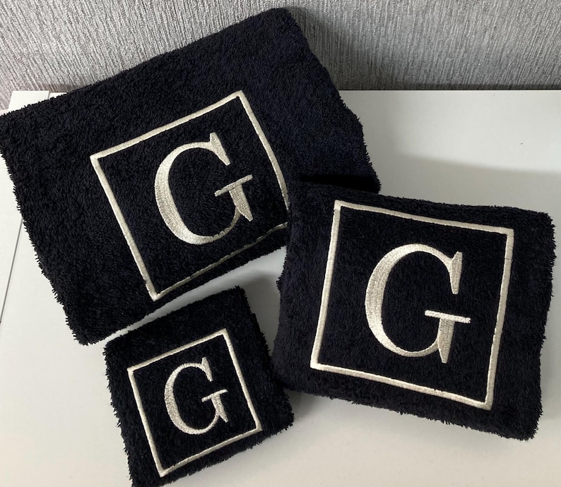 May include: A set of three black terry cloth towels. Each towel features a white embroidered square with the letter "G" in the center. The towels are in varying sizes, suitable for a bathroom or spa.