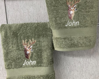 May include: Two olive green terry cloth towels hanging on a metal rack. Each towel features an embroidered deer head with antlers and the name John stitched in white thread above a horizontal chevron-patterned band near the bottom edge.
