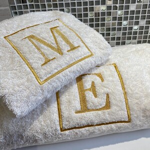 May include: Two white bath towels with gold embroidered initials, "M" and "E", within a gold square border. The towels are fluffy and appear to be made of a soft material. The towels are folded and stacked on top of each other.