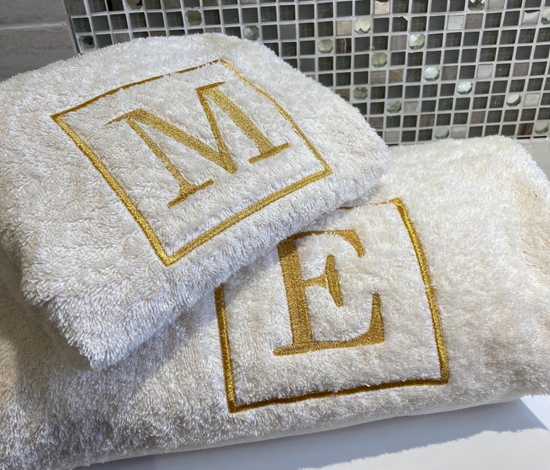 May include: Two white bath towels with gold embroidered initials, "M" and "E", within a gold square border. The towels are fluffy and appear to be made of a soft material. The towels are folded and stacked on top of each other.
