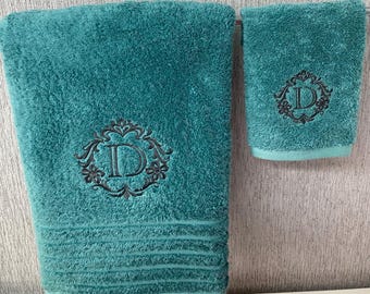 May include: Teal bath towel and hand towel set. Both towels feature an embroidered black letter "D" within a decorative floral frame. The bath towel has horizontal ribbed detailing near the bottom.