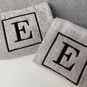 May include: Two gray washcloths with a black embroidered letter "E" inside a black square. The washcloths are made of a soft, absorbent material and are ideal for use in the bathroom or kitchen.
