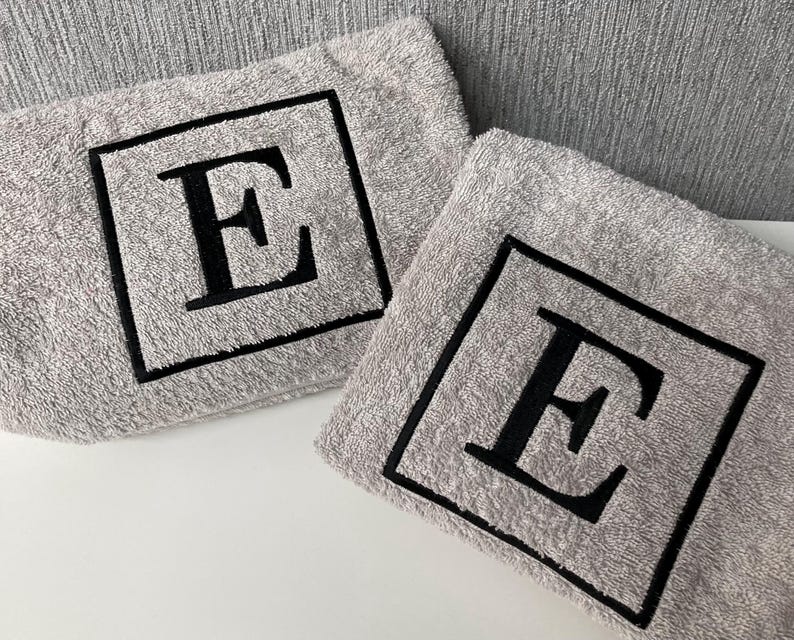 May include: Two gray washcloths with a black embroidered letter "E" inside a black square. The washcloths are made of a soft, absorbent material and are ideal for use in the bathroom or kitchen.
