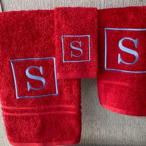 May include: A set of three red bath towels with a light blue embroidered letter "S" inside a square frame. The towels vary in size, with a bath towel, a hand towel, and a washcloth. The towels are made of a soft, absorbent material.