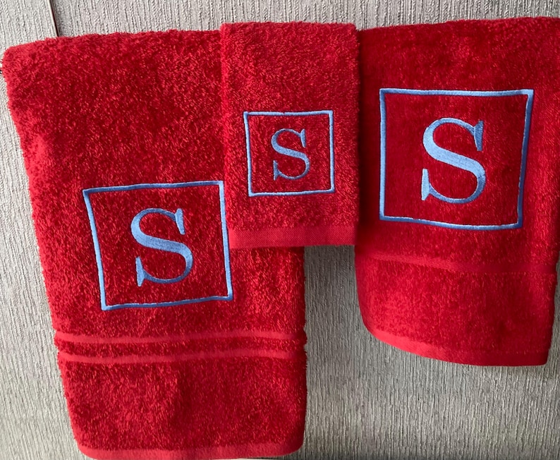 May include: A set of three red bath towels with a light blue embroidered letter "S" inside a square frame. The towels vary in size, with a bath towel, a hand towel, and a washcloth. The towels are made of a soft, absorbent material.