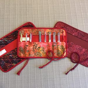 May include: A red and orange batik fabric knitting needle roll with several knitting needles inside. The roll is open, displaying the needles, and is accompanied by two matching red fabric cases. The cases and roll are tied with red cords.