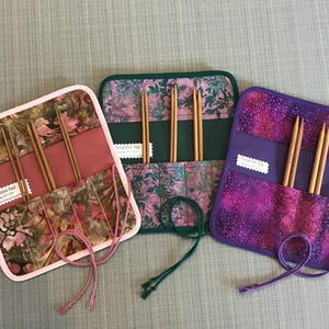Può includere: Tre custodie per ferri da maglia in tessuto rosa, verde e viola, ciascuna con ferri da maglia in legno. Le custodie sono rettangolari con chiusura a patta e un cordino abbinato. Il tessuto ha un motivo floreale.