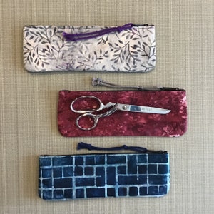 May include: Three fabric zippered pouches with different patterns. The top pouch is a light gray with a floral pattern. The middle pouch is a dark red with a floral pattern. The bottom pouch is a dark blue with a geometric pattern.