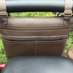 May include: A brown, faux leather bicycle handlebar bag with a zippered front pocket and two horizontal stitched lines. The bag has brown nylon straps with gold-colored metal buckles. The bag is attached to a black handlebar.