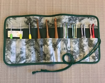 Puede incluir: Un estuche enrollable de tela verde con cierre de corbata, que contiene una variedad de ganchos de ganchillo. El estuche tiene un patrón de mármol blanco y gris. El texto "Kingfisher Bags" es visible en una pequeña etiqueta.