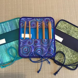 Puede incluir: Tres estuches para agujas de tejido diferentes con patrones azules, verdes y morados. Cada estuche tiene una correa azul y una etiqueta que dice "Knitting Needle Case".