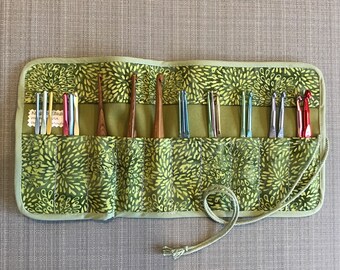 Puede incluir: Un estuche enrollable de tela verde con un patrón floral y un cierre de cordón. El estuche tiene múltiples ranuras para guardar ganchos de ganchillo. Los ganchos son de madera, metal y plástico.